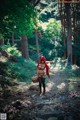 A woman in a red riding hoodie is walking through the woods.