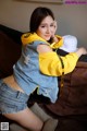 A woman sitting on a couch wearing a yellow hoodie and denim shorts.